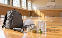 中学校の体育館の床に並べられた非常用持ち出し袋と、カエルの小さな置物