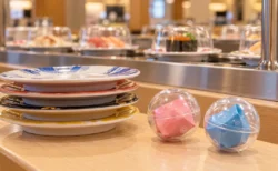 回転寿司のカウンターに置かれた寿司皿と、小さなカプセルトイの景品が入った透明カプセル