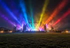 夜の野外フェス会場で、虹色のステージ照明が夜空へ放射状に伸びる様子