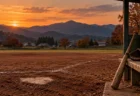 秋の夕暮れ、山並みを望む町の野球場。土のグラウンドのホームベースと、ベンチに立てかけた木製バットと革のグローブ