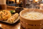 木のカウンターに置かれた湯気の立つ釜揚げうどんの桶と、かき揚げ・ちくわ天の皿