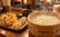 木のカウンターに置かれた湯気の立つ釜揚げうどんの桶と、かき揚げ・ちくわ天の皿