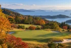 秋の紅葉に囲まれたゴルフ場のグリーンと黄色いピンフラッグ、遠くに島が浮かぶ海