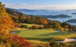 秋の紅葉に囲まれたゴルフ場のグリーンと黄色いピンフラッグ、遠くに島が浮かぶ海
