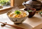 料亭の白木のカウンターに置かれた湯気の立つ丼と木の箸、奥に土鍋と吸い物の椀