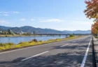 秋の日差しの中、川沿いに続く道路と色づく木々