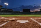 ナイター照明が灯る野球場の本塁ベースと、そばに置かれた木製バットのイメージ