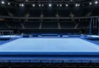 照明に照らされた体操競技会場の青いゆかのフロアと、奥に並ぶ平行棒やつり輪の器具