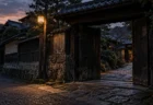 夕暮れの古い住宅街に立つ重い木の門と、その先に続く石畳の道