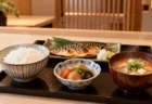木のカウンターに並ぶ炊きたての白いご飯と味噌汁、焼き魚の和食膳