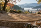 朝の斜光が差す屋外の稽古土俵と、俵のそばに置かれた塩かごと竹ぼうき
