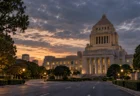 夕暮れの空の下に建つ重厚な石造りの議事堂風の建物と、手前の並木道