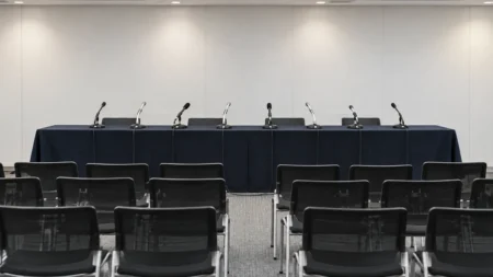 長机にマイクが並び、手前に無人の椅子が並ぶ記者会見場