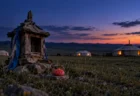 夕暮れの草原に並ぶ白いゲルと、手前の小さな木の祠に供えられた赤い饅頭