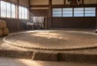 朝の光が差し込む相撲部屋の稽古場の土俵と俵、壁際に掛けられた白い稽古まわし