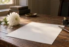 朝の光が差す木の机に置かれた白い菊とつけペン、インク瓶、白い原稿用紙